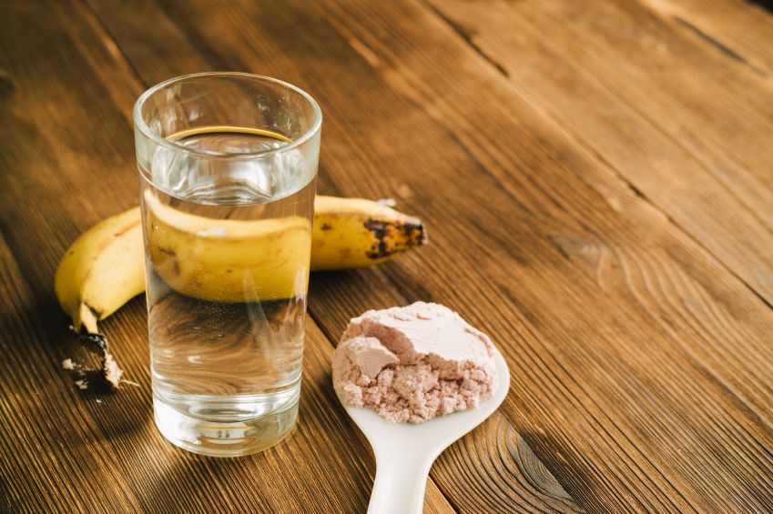 Sports protein on the spoon, a banana and a glass of water on wooden table