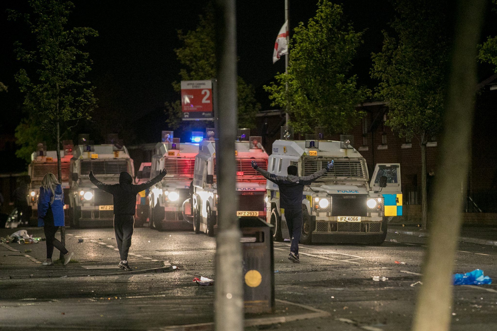Two men taunt police officers in their riot vans, spreading their arms wide above their heads, a flag bearing the Red Hand of Ulster flies in the background